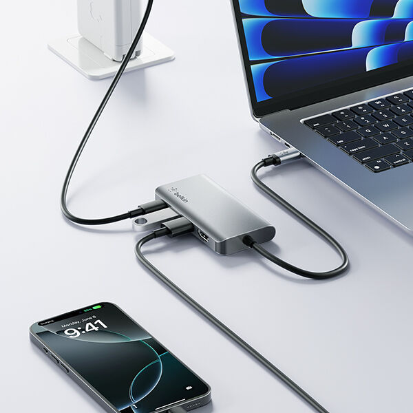 A sleek and modern workspace featuring a MacBook connected to a Belkin USB-C hub. The hub is actively connecting multiple devices, including a smartphone and an external display, with neatly arranged cables on a clean white desk. A wireless charger with an Apple device is visible in the background.