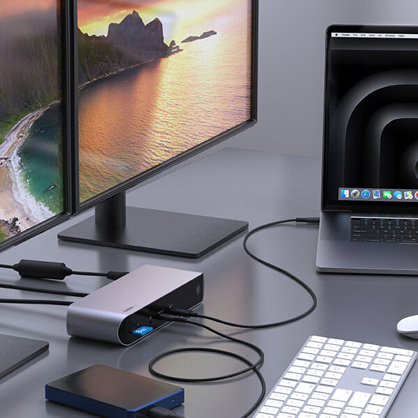 A Belkin docking station connected to a laptop, external hard drive, and dual monitors displaying a scenic sunset view, with a keyboard and mouse on the desk.