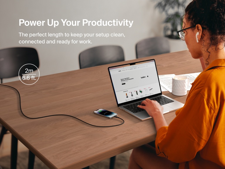 A woman works at a desk with her iPhone charging via a 2-meter (6.6 ft) Belkin BoostCharge Braided USB-A Cable with Lightning Connector, ideal for maintaining a clean, connected workspace.