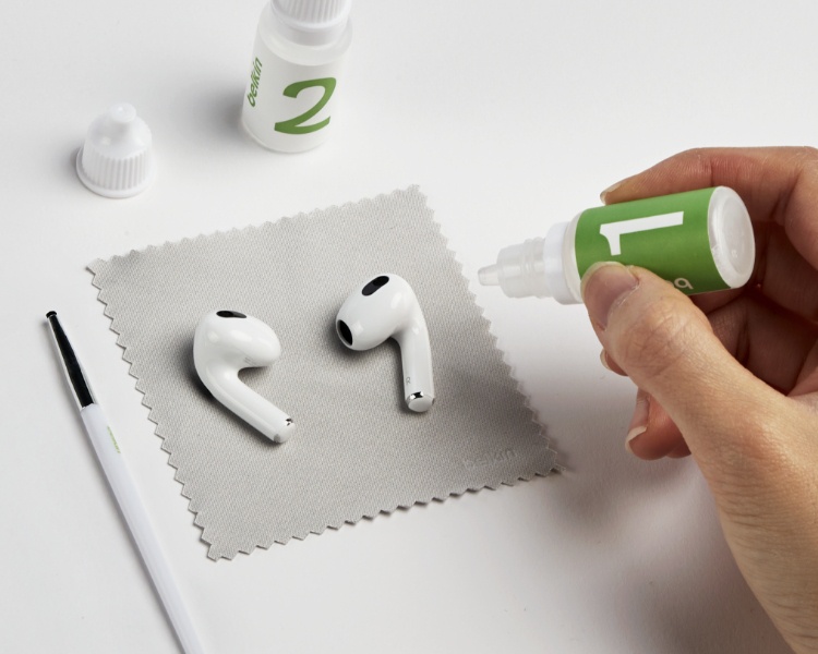 The image displays a cleaning setup for earbuds. Two white wireless earbuds are placed on a soft cleaning cloth, with a cleaning solution bottle labeled "1" being held and applied by a hand. Another bottle labeled "2" and a cleaning tool or pen are also visible on the white surface. The arrangement emphasizes a step-by-step cleaning process for maintaining earbuds.