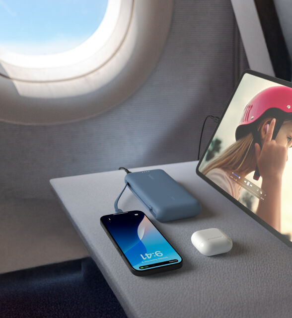 A blue Belkin BoostCharge Power Bank 20K with Integrated Cable is placed on an airplane tray table, charging an iPhone and iPad, with a pair of AirPods nearby. A blue Belkin BoostCharge Power Bank 20K with Integrated Cable is placed on an airplane tray table, charging an iPhone and iPad, with a pair of AirPods nearby.