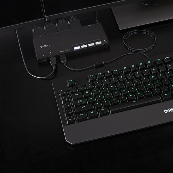Belkin KVM switch connected to a keyboard and multiple cables on a dark desk, shown from an overhead angle with illuminated keyboard keys.