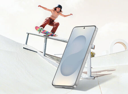 Smartphone standing upright in a skate park foreground while a skateboarder performs a trick on a rail behind it, suggesting durability in active environments.