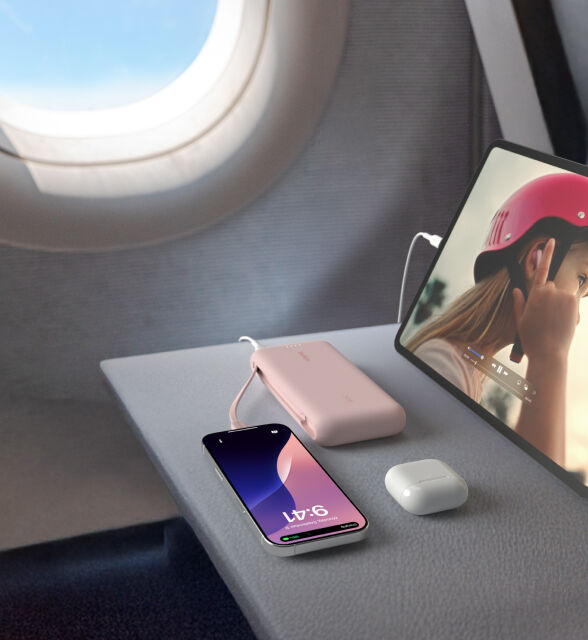 A pink Belkin BoostCharge Power Bank 20K with Integrated Cable is placed on an airplane tray table, charging an iPhone and iPad, with a pair of AirPods nearby. A pink Belkin BoostCharge Power Bank 20K with Integrated Cable is placed on an airplane tray table, charging an iPhone and iPad, with a pair of AirPods nearby.