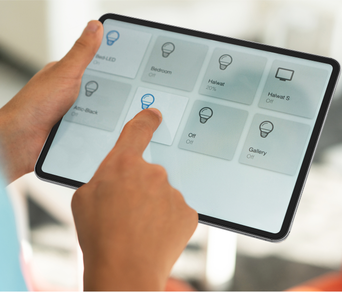 Primer plano de una persona que utiliza una tableta para controlar la iluminación de su hogar inteligente. La pantalla muestra varias opciones de control de iluminación para diferentes habitaciones, con íconos que indican el estado de cada luz. El usuario toca una de las opciones de iluminación, lo que demuestra la facilidad de ajuste de la configuración a través de la interfaz de pantalla táctil.