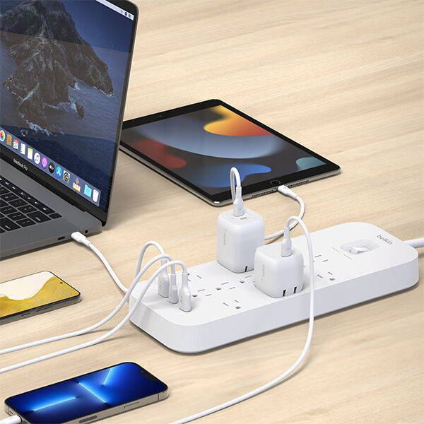 A white power strip on a wooden desk with multiple charging adapters connected, powering a laptop, tablet, and several smartphones at the same time.