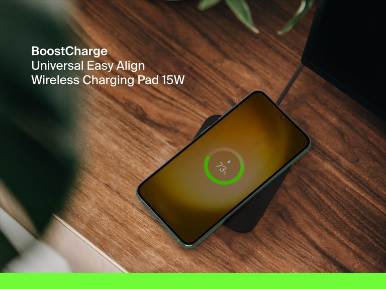 A smartphone charges on the Belkin BoostCharge Universal Easy Align Wireless Charging Pad 15W, shown on a wooden desk with an on-screen indicator displaying 73% battery.