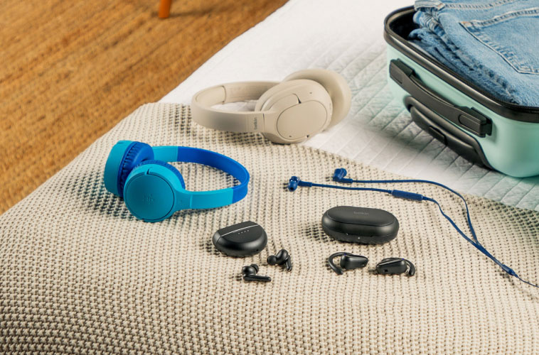 A variety of Belkin headphones and earbuds—including over-ear, on-ear, wireless, and wired models—neatly arranged on a bed next to an open mint green suitcase packed with clothes.