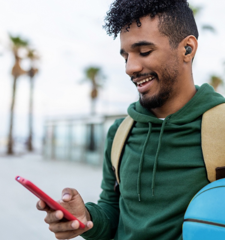 A young man wearing a green hoodie, beige backpack, and wireless earbuds smiles while looking at his smartphone. He is holding a red phone and standing outdoors, with palm trees and a clear sky in the background. A blue basketball is partially visible in the lower right corner.