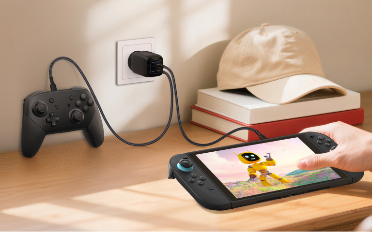 Een Nintendo Switch 2 en een gamecontroller worden opgeladen op een houten bureau,bij een zonnig raam. De console wordt vastgehouden door een persoon en toont een heldere, vrolijke scène met een gele robot in een kleurrijk veld. Beide apparaten zijn met een zwarte USB-laadkabel aangesloten op een wandadapter die in een stopcontact is gestoken. In de buurt ligt een beige baseballpet op een stapel boeken, wat zorgt voor een gezellige en netjes ingerichte plek om te gamen.
