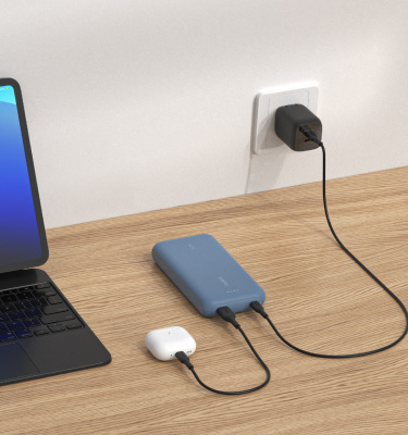 파란색 케이블 일체형 Belkin BoostCharge 보조 배터리 20K가 나무 표면 위에 놓인 상태로 AirPods을 충전하는 동시에 벽면 콘센트를 통해 충전되고 있고, 태블릿이 근처에 놓여 있습니다.