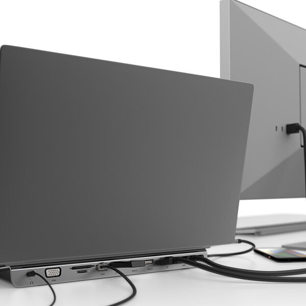 A close-up view of the back of a laptop connected to a docking station with multiple ports, including VGA, HDMI, USB, and Ethernet. The docking station is neatly integrated with the laptop, managing various cables that lead to an external monitor and other peripherals. The workspace is clean and modern, emphasizing efficient cable management and connectivity.
