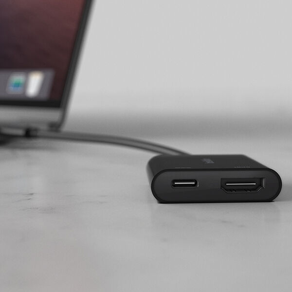  A close-up of a black USB-C hub connected to a laptop on a white marble surface. The hub features a USB-C port and an HDMI port, providing additional connectivity options for the laptop. The blurred background showcases part of the laptop screen and keyboard.