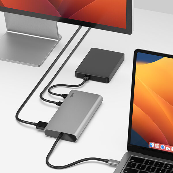 A modern workspace setup featuring a laptop connected to an external monitor, external hard drive, and other peripherals through a sleek, silver Belkin docking station. The docking station helps streamline cable management, supporting multiple connections via USB-C, HDMI, and additional ports for enhanced productivity.