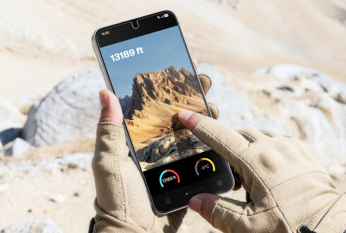 Gloved hands holding a smartphone outdoors, displaying a mountain landscape on the screen with an altitude reading of 13,189 feet.