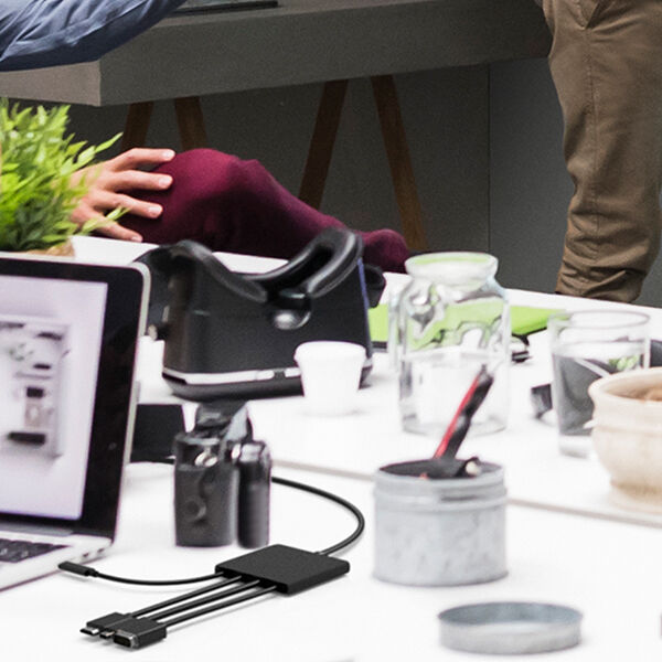 A modern office workspace with a white table cluttered with various items, including a laptop displaying a webpage, a digital camera, a black multi-port hub with connected cables, glass jars, a small potted plant, and a coffee cup. In the background, people are engaged in conversation, with one person gesturing while another listens. The setting appears to be a collaborative workspace or meeting area.