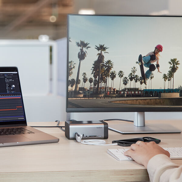 Creative workspace with a laptop connected to a monitor via a Belkin docking station, showcasing video editing on the laptop and a skateboarder in action on the monitor.