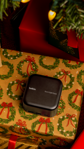 Ein Belkin Connect USB-C-6-in-1-Core-GaN-Dock (130 W) ist auf einem Geschenkkarton unter einem Weihnachtsbaum zu sehen, der von Weihnachtsdekoration umgeben ist.