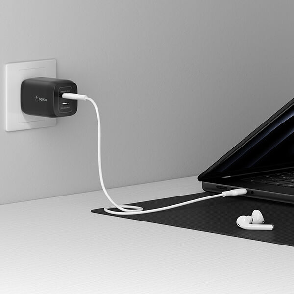 A black Belkin wall charger plugged into an outlet, with a white charging cable running to a laptop on a desk, and a pair of wireless earbuds resting nearby on a desk mat.