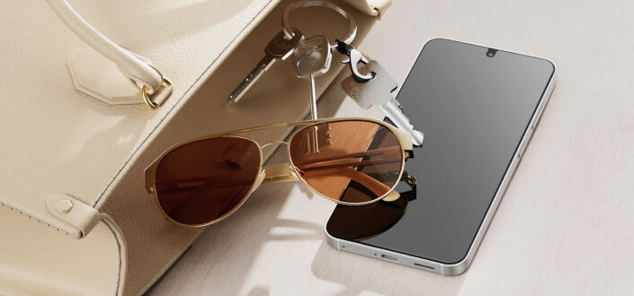 A smartphone resting on a light-colored surface beside a beige handbag, sunglasses, and keys, suggesting everyday carry and lifestyle use.
