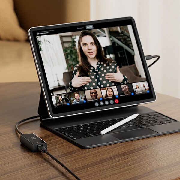 A tablet with a detachable keyboard on a wooden table, displaying a video conference call with multiple participants. A white stylus rests on the keyboard, and a black Belkin adapter is connected to the tablet via a USB-C cable. The background features a cozy indoor setting with neutral tones.