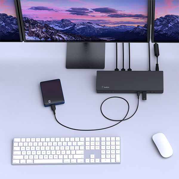 Overhead view of a minimalist desk setup with a widescreen monitor displaying a mountain landscape, a docking station with multiple cables connected, an external hard drive, a white keyboard, and a white mouse arranged neatly on a light-colored desk.