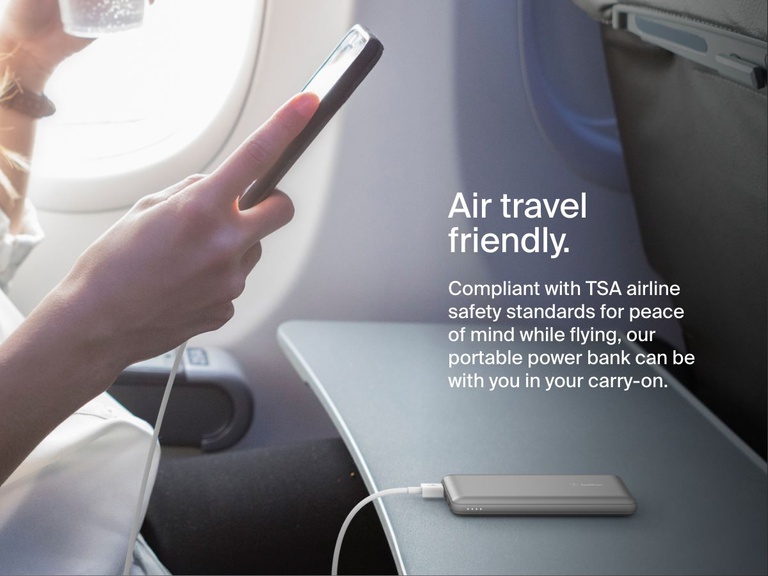 A person uses a smartphone on an airplane while a silver Belkin BoostCharge Power Bank 10K rests on the tray table, illustrating its TSA-compliant, travel-friendly design for carry-on use.