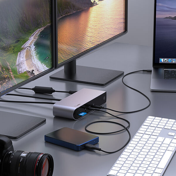 A professional workspace setup featuring a laptop connected to dual external monitors through a Belkin docking station. The docking station also connects to an external hard drive, keyboard, and other peripherals, creating an organized and efficient workstation. A camera is placed on the desk, suggesting a setup optimized for creative professionals or content creators.