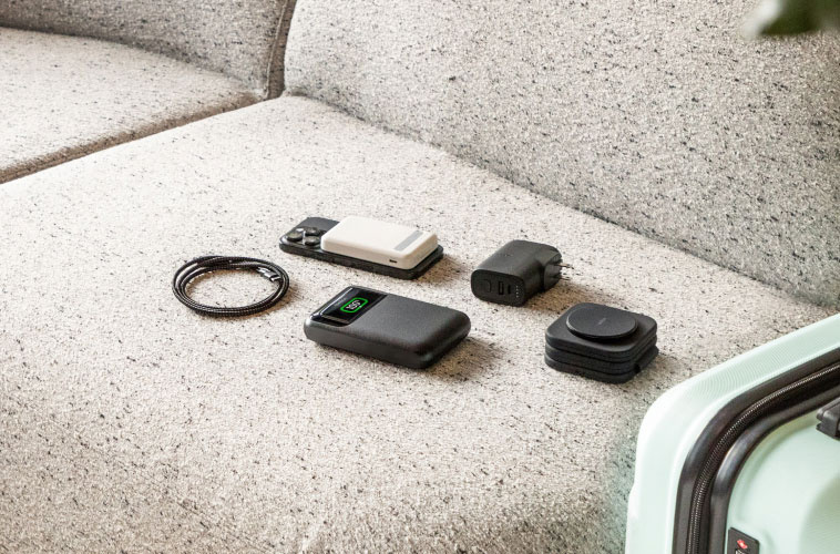 Flat lay of MagSafe portable charger, USB-C cables, and travel adapters on hotel bench next to suitcase