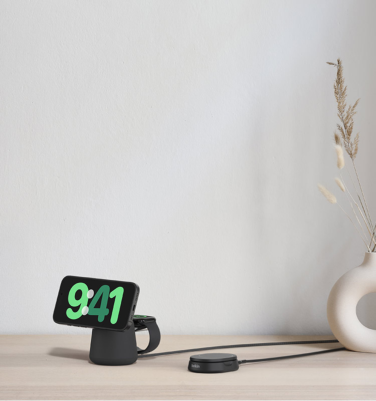 A black Belkin Convertible Magnetic Wireless Charging Stand with Qi2 15W and a Belkin 2-in-1 Wireless Charging Dock with MagSafe 15W in Charcoal are displayed on a light wood surface next to a white vase with dried grasses.