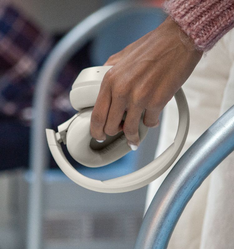 Close-up image of a hand holding a pair of folded sand over-ear headphones near a metallic rail, with a blurred background including another person seated in the distance.