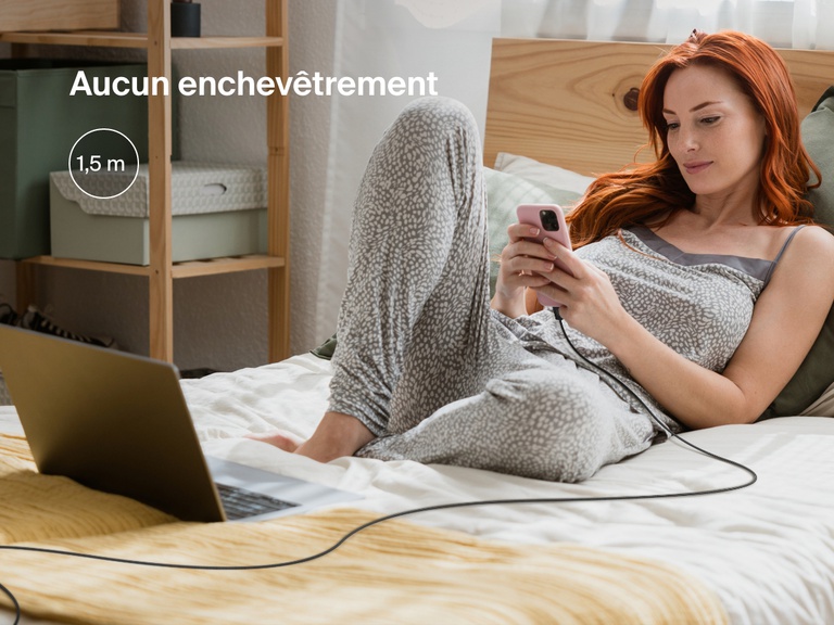 Une femme confortablement installée sur un lit utilise son iPhone connecté à un câble USB-C Belkin BoostCharge avec connecteur Lightning de 1,8 m de long pour une recharge pratique, même loin d'une prise.