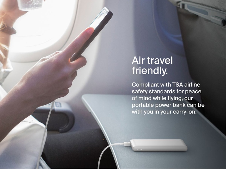 A person uses a smartphone on an airplane while a white Belkin BoostCharge Power Bank 10K rests on the tray table, illustrating its TSA-compliant, travel-friendly design for carry-on use.