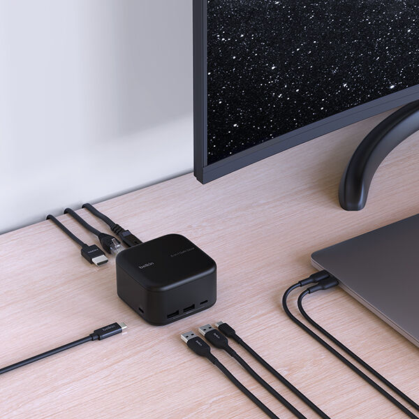 A sleek and organized workspace featuring a Belkin docking station connected to a laptop and an external monitor. Various cables, including HDMI and USB, are neatly arranged, demonstrating the docking station's multiple connectivity options. The monitor displays a starry night sky, and the setup is placed on a clean wooden desk, creating a modern and efficient workstation.