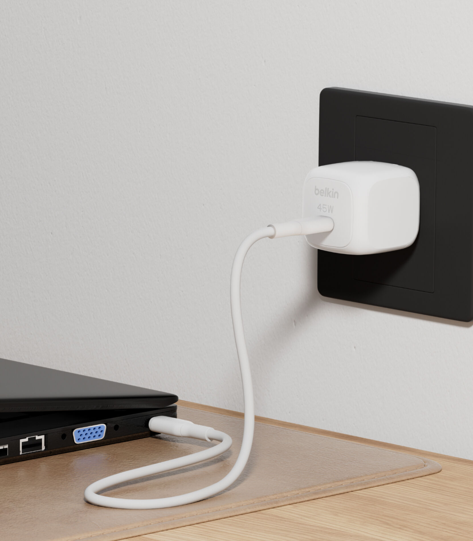 Belkin BoostCharge Cubic Charger 45Wを壁の黒色のコンセントに差し込み、USB-Cから白色のUSB-Cケーブルを使って木製の机の上のノートパソコンを充電しています。