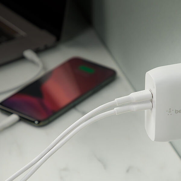 Close-up of a white wall charger with two cables plugged in, charging a smartphone lying on a desk, with part of a laptop visible in the background.
