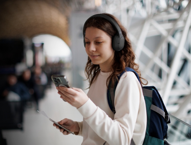 画像には、オーバーイヤーヘッドホンを装着し、バックパックを背負った若い女性が写っています。彼女は片手に持ったスマートフォンを見ながら、わずかに微笑んでいます。彼女が写る背景はぼやけた明るいモダンな屋内環境で、空港や駅のような公共の場である可能性を示唆しています。