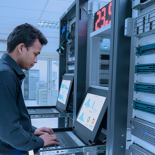 A technician working in a server room, monitoring data displayed on screens mounted in server racks. The screens show graphs and charts, while a digital temperature display above reads 23.9°C. Networking equipment and cabling are organized in the racks beside him, highlighting a controlled and well-maintained environment.