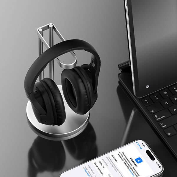 A pair of black over-ear headphones resting on a sleek silver headphone stand, placed on a reflective black desk next to a tablet with a keyboard and a smartphone displaying Bluetooth settings.