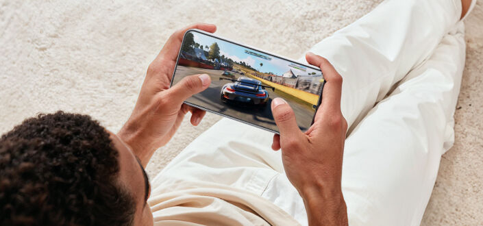 A person holding a smartphone in landscape mode while playing a racing video game, with a car driving on a track displayed on the screen.