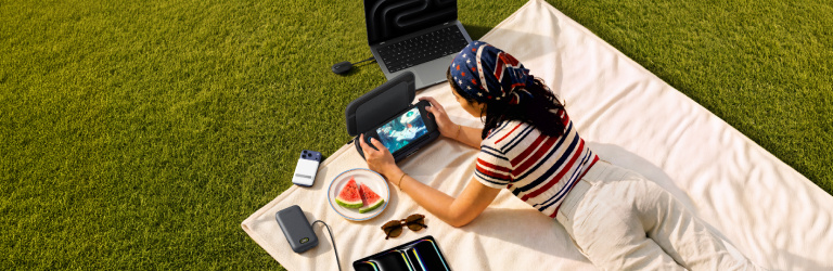 Woman relaxing outdoors on a picnic blanket while gaming on a handheld device surrounded by Belkin tech accessories and a laptop.