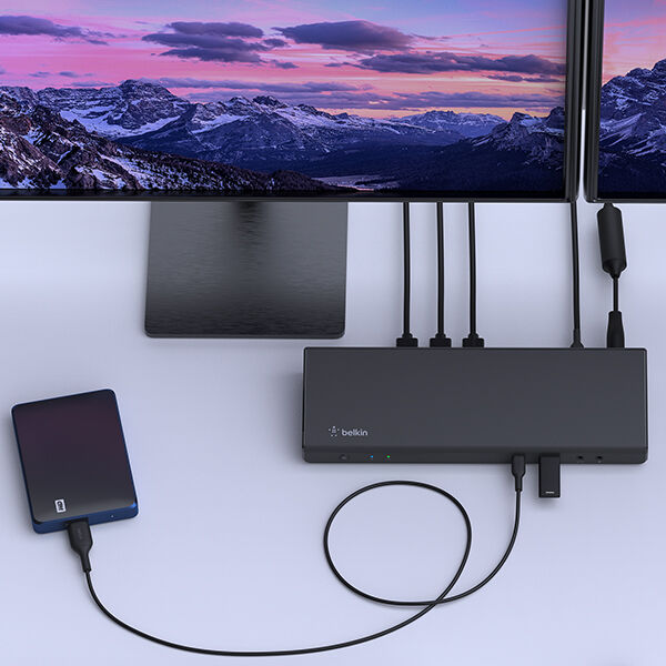 A sleek Belkin docking station on a desk, connected to two high-resolution monitors displaying a scenic mountain landscape. Multiple cables are plugged into the docking station, including HDMI and USB connections. An external hard drive is also connected via a USB cable, demonstrating an organized and efficient workstation setup.