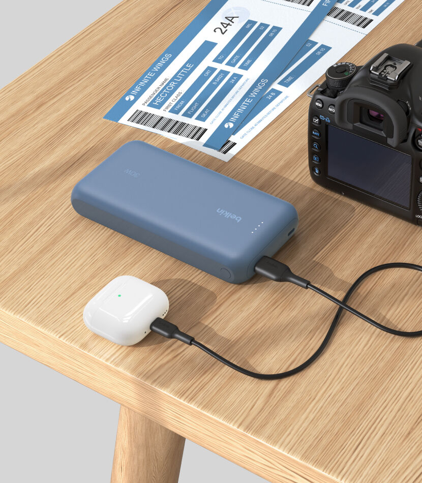 A blue Belkin BoostCharge Power Bank 20K with Integrated Cable is placed on a wooden table, charging a pair of white AirPods, with a DSLR camera and boarding passes nearby. A blue Belkin BoostCharge Power Bank 20K with Integrated Cable is placed on a wooden table, charging a pair of white AirPods, with a DSLR camera and boarding passes nearby.