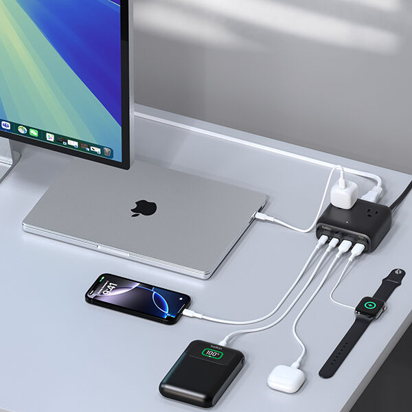 A desk setup with a laptop, smartphone, wireless earbuds, smartwatch, and a portable battery pack all charging simultaneously from a compact multi-port power hub beside a monitor.