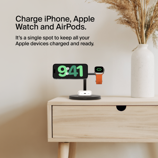 This image displays a sleek charging stand designed to charge an iPhone, Apple Watch, and AirPods simultaneously. The devices are neatly arranged on the stand, which sits atop a light wooden nightstand. The iPhone screen shows the time as 9:41, the Apple Watch features a green charging indicator, and the AirPods case is positioned on the base of the stand. The text emphasizes the convenience of "a single spot to keep all your Apple devices charged and ready." A decorative vase with dried plants complements the minimalistic and organized setup.