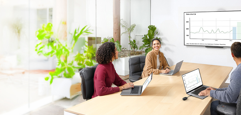 Three people sit around a conference table with laptops, smiling and discussing a presentation. A large wall-mounted screen displays a line graph labeled &lsquo;Data chart.&rsquo; The room has glass walls, natural light, and several green plants, creating a bright, modern meeting space.