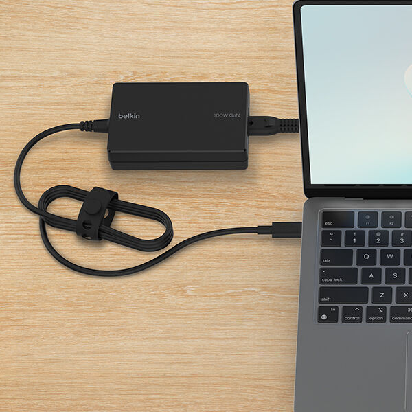 A Belkin 100W GaN charger connected to a MacBook via a USB-C cable, neatly coiled and secured with a cable strap, placed on a wooden surface.