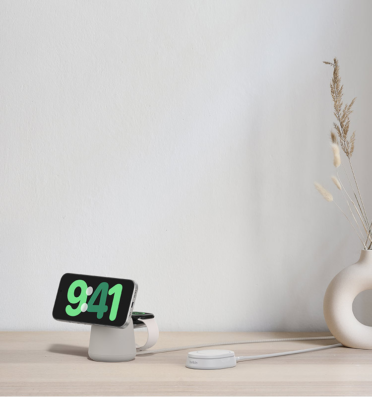 A white Belkin Convertible Magnetic Wireless Charging Stand with Qi2 15W and a Belkin 2-in-1 Wireless Charging Dock with MagSafe 15W in Sand are displayed on a light wood surface next to a white vase with dried grasses.