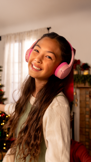 A smiling young girl wears Belkin SoundForm Mini Wireless On-Ear Headphones for Kids in a festive, holiday-themed living room, with a decorated Christmas tree and fireplace in the background.
