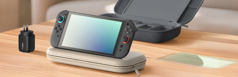 A Nintendo Switch 2 console is propped on a light-colored protective carrying case atop a wooden desk. Nearby, a black wall charger stands upright, and an open dark gray storage case with compartments is visible in the background. A screen protector lies flat on the desk, completing the setup of essential gaming accessories in a clean, well-lit room.
